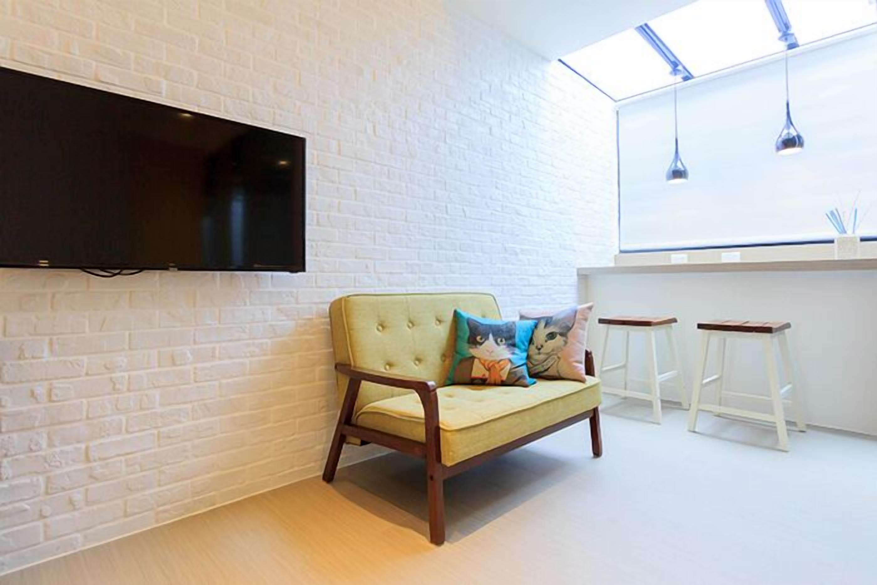 A modern room with a white brick wall, a flat-screen TV mounted above a light yellow sofa with cat-themed cushions, and a white bar counter with stools.