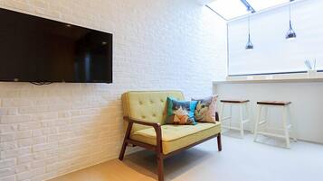 A modern room with a white brick wall, a flat-screen TV mounted above a light yellow sofa with cat-themed cushions, and a white bar counter with stools.