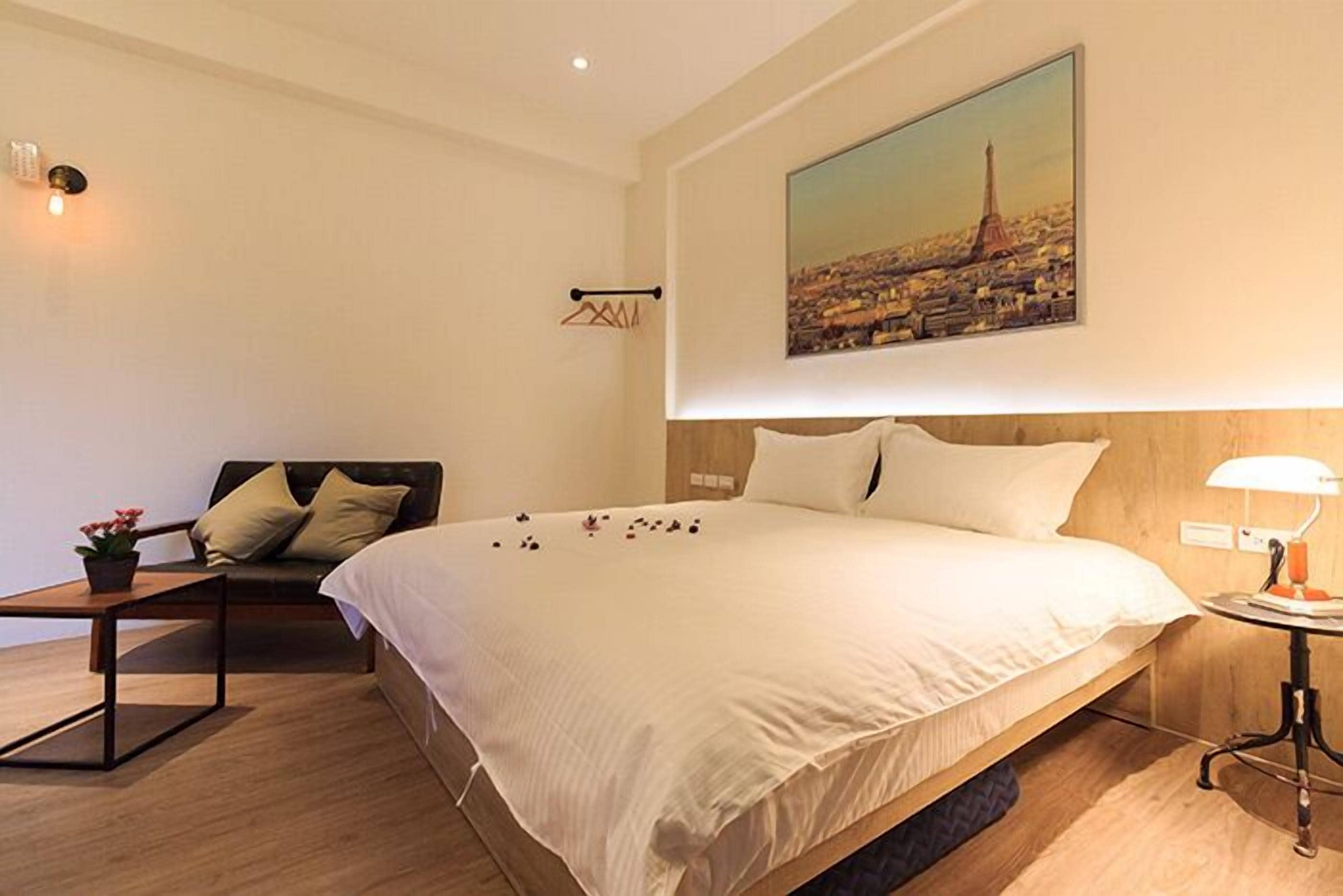Une chambre d’hôtel avec un grand lit, une table de chevet avec une lampe, une petite table avec une plante et un tableau représentant un paysage urbain accroché au mur.