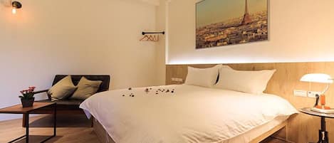 Een hotelkamer met een groot bed, een nachtkastje met een lamp, een kleine tafel met een plant, en een schilderij van een stadsgezicht aan de muur.