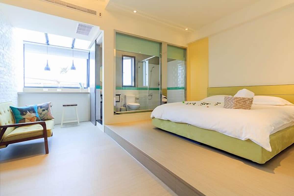 Un dormitorio moderno con una cama grande, un rincón de descanso pequeño y un baño visible a través de una puerta de cristal.