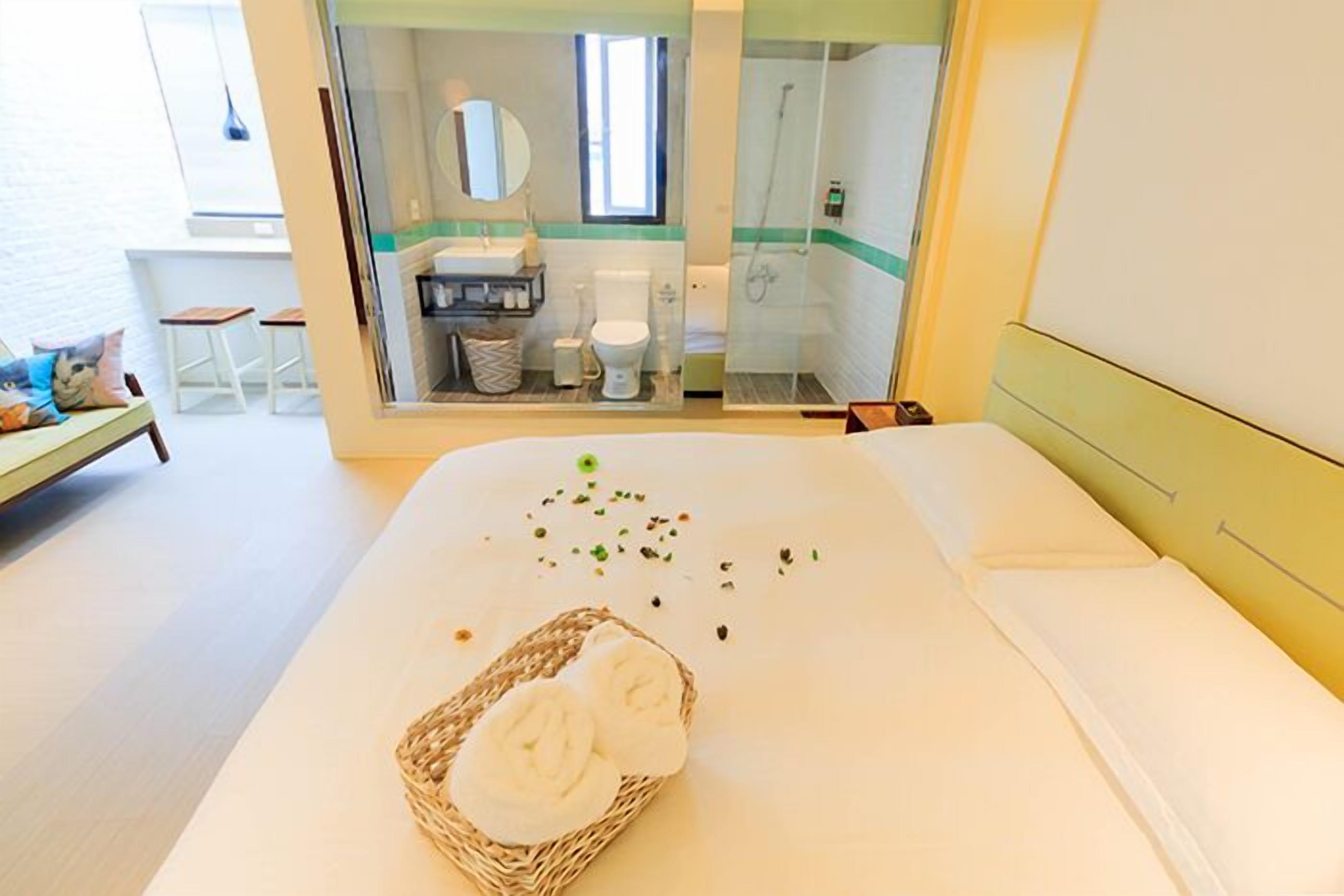 Une chambre d’hôtel avec un lit, un panier en osier contenant des serviettes, une salle de bain visible à travers une porte en verre et une vue sur la mer.