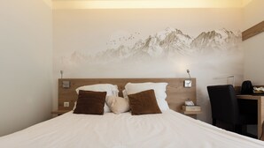 Een bed met witte lakens en bruine kussens, een houten hoofdbord, een nachtkastje met een lamp en een bureau met een stoel.