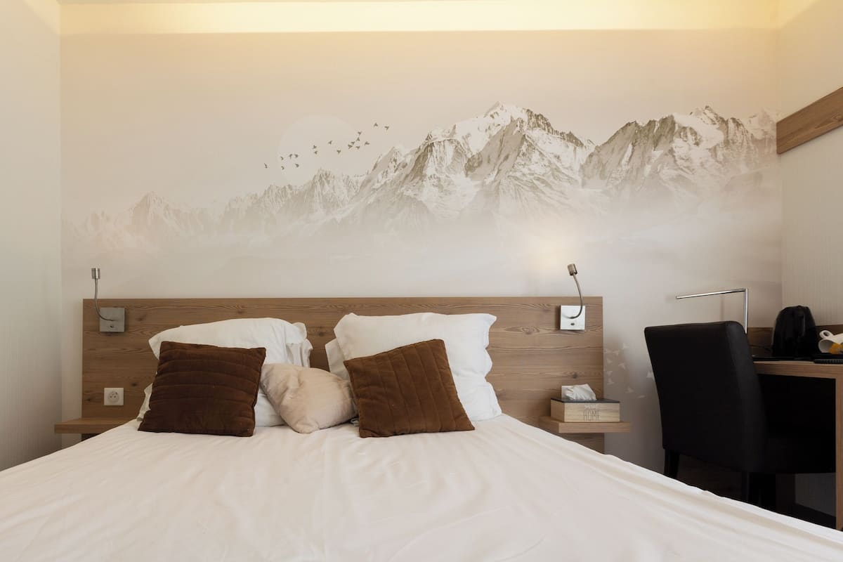Een bed met witte lakens en bruine kussens, een houten hoofdbord, een nachtkastje met een lamp en een bureau met een stoel.