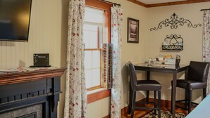 A room with a fireplace, a desk with chairs, a television, and a window with curtains.