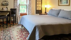 A bedroom with a large bed, a desk with a chair, a window with curtains, a lamp, and a framed picture on the wall.