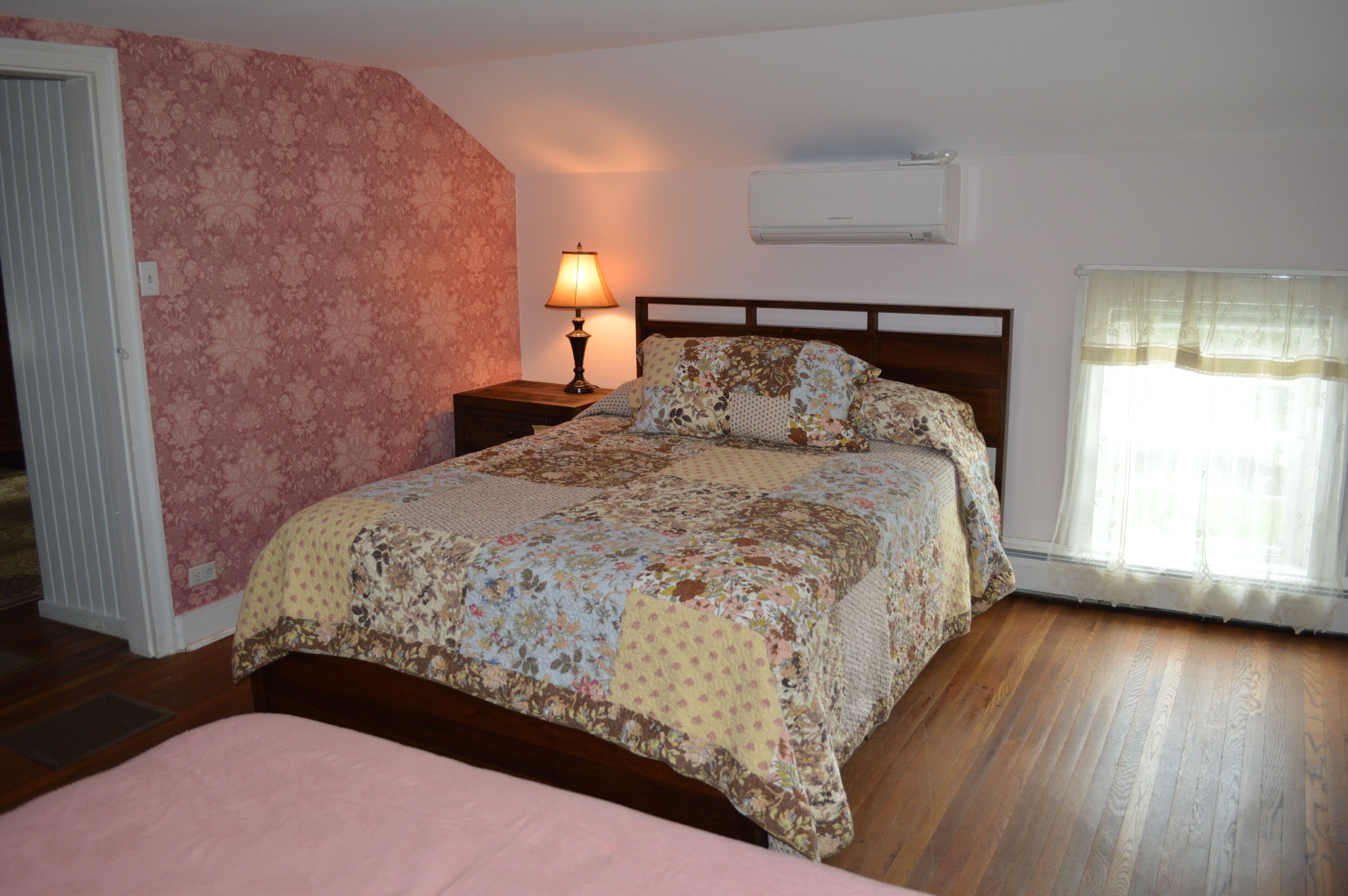 Un dormitorio con edredón estampado con flores, una lámpara de noche, un cabecero de madera y una ventana con cortinas translúcidas.