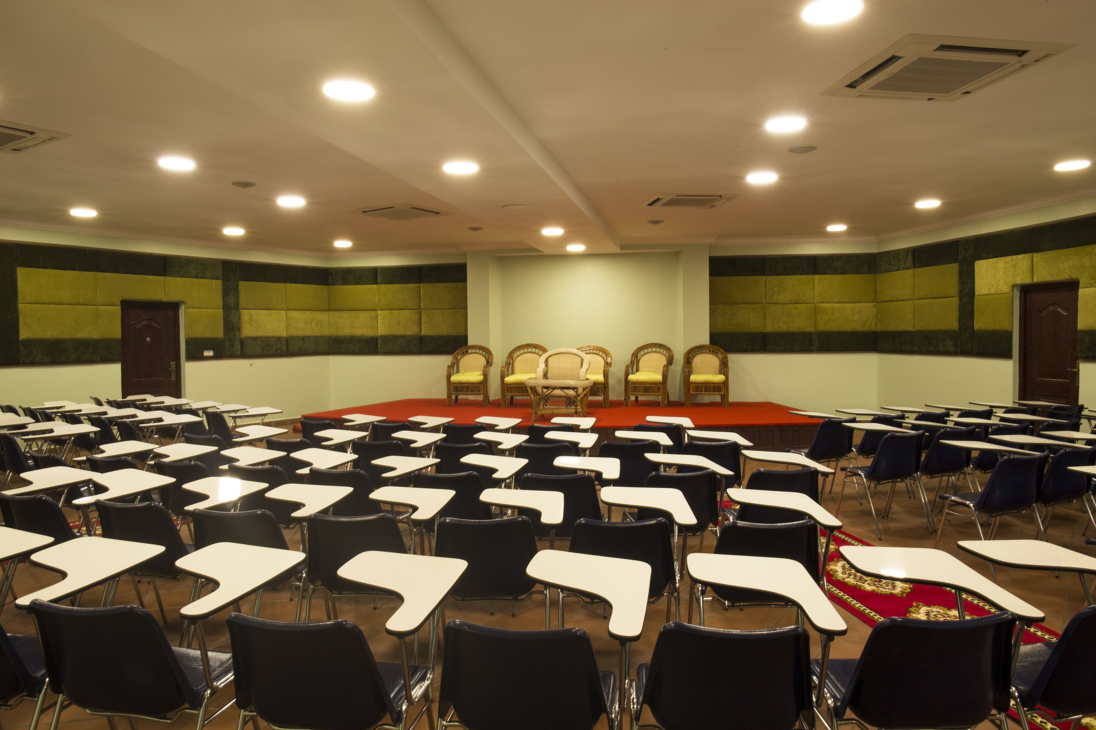 A conference room with rows of desks and chairs, a stage with a podium, and a carpeted floor.