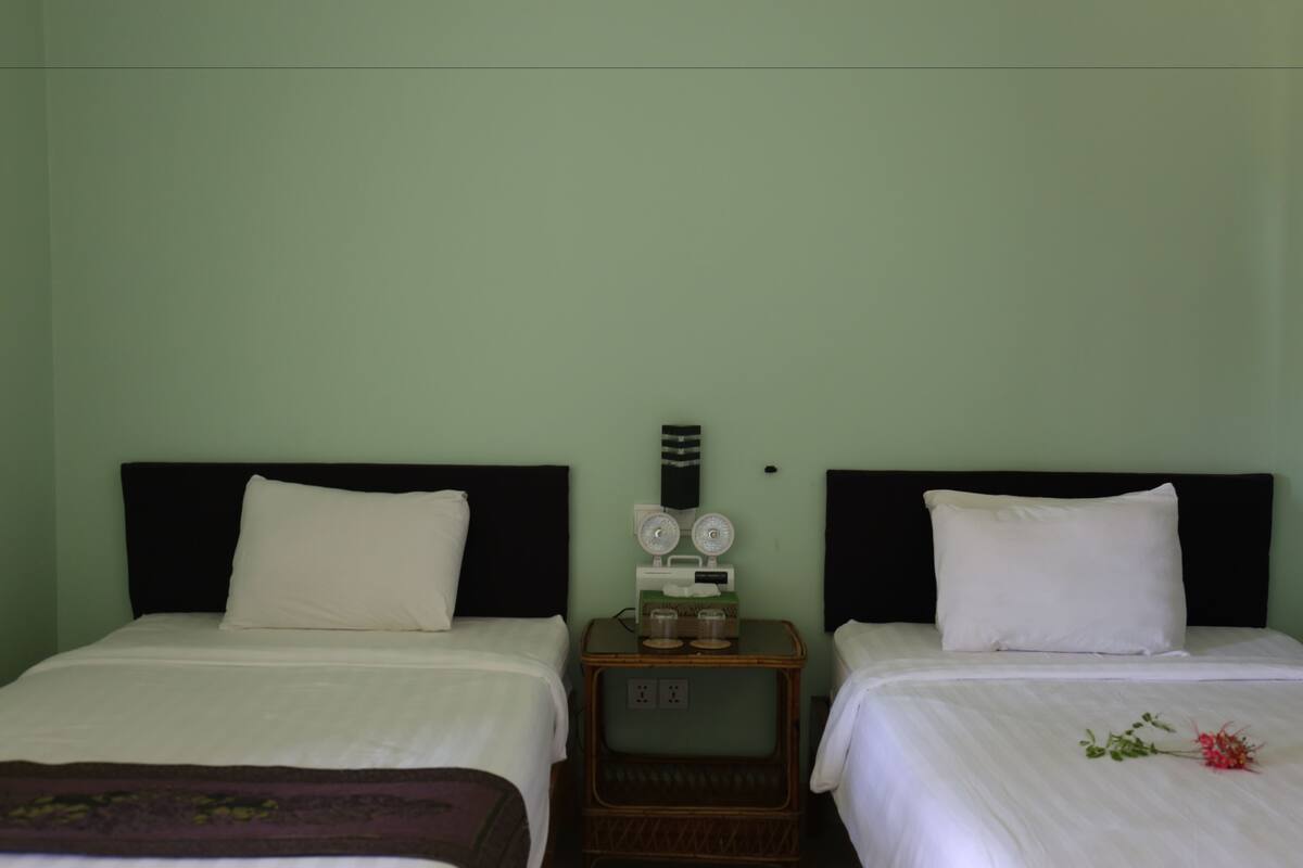 Habitación de hotel con dos camas, cada una con ropa blanca y cabeceros negros. Una pequeña mesita de madera entre las camas tiene un reloj y un enchufe.