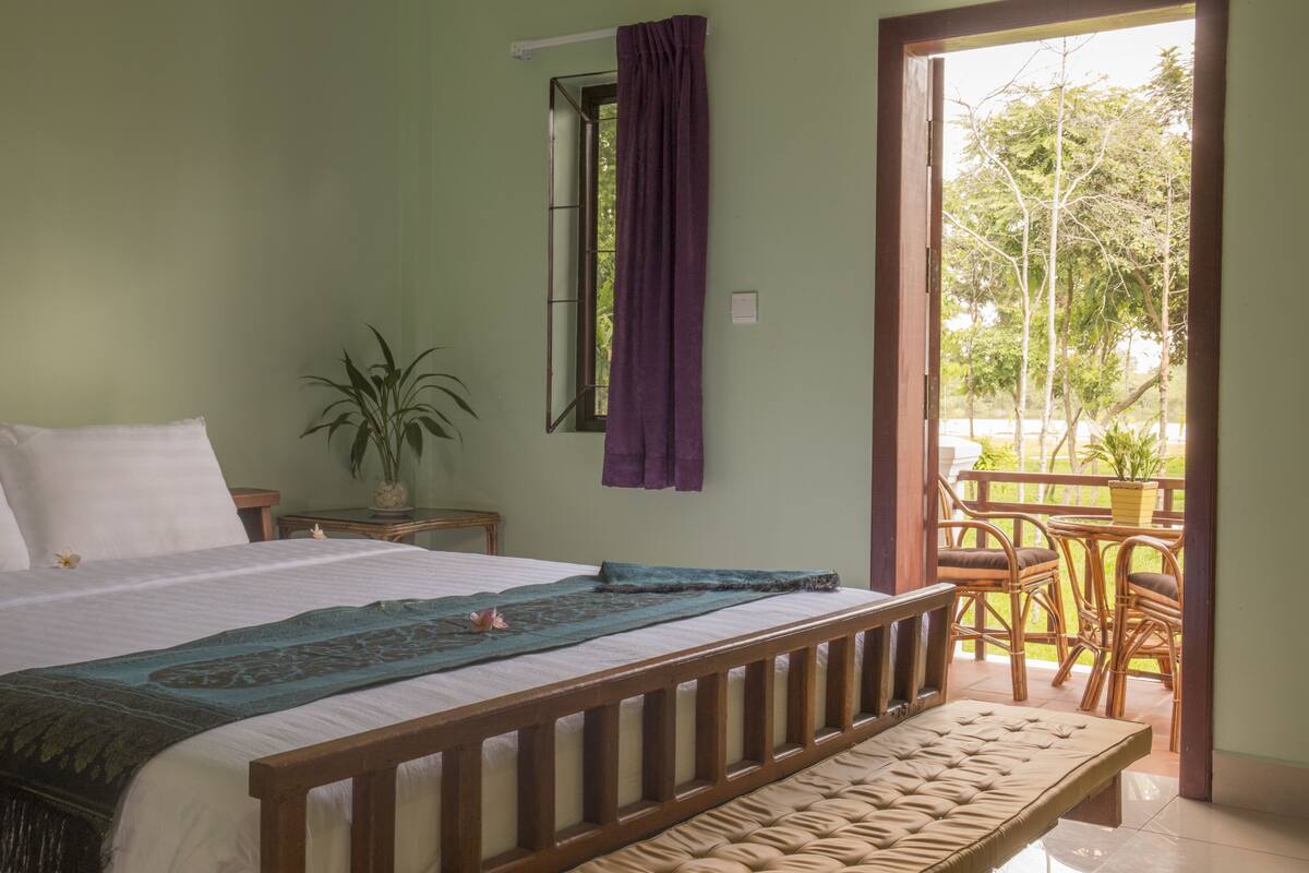 Un dormitorio con cama, ventana con cortinas moradas, mesita de noche con planta y balcón con muebles de mimbre.