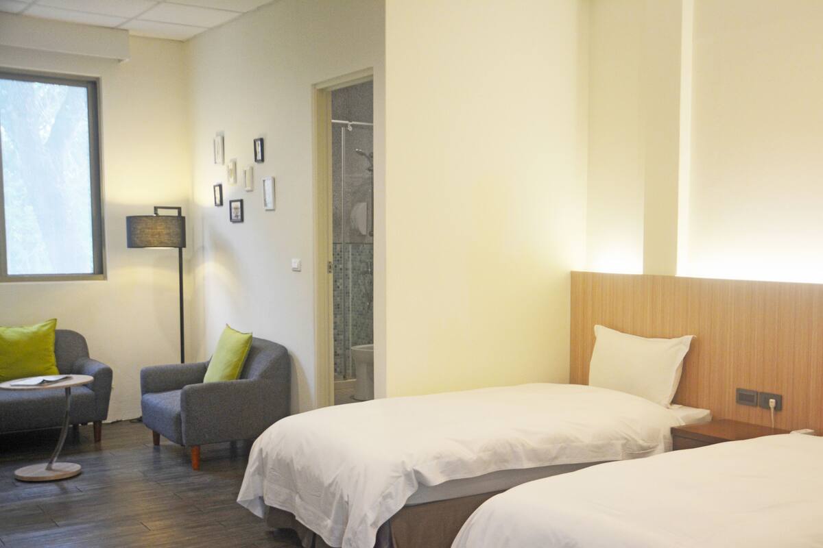 A hotel room with two beds, a sofa, a small table, and a bathroom with a glass shower.
