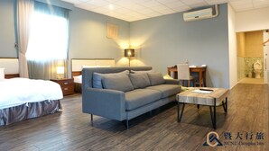 Habitación de hotel con cama, sofá gris, una mesita, escritorio y baño visible a través de una puerta abierta.