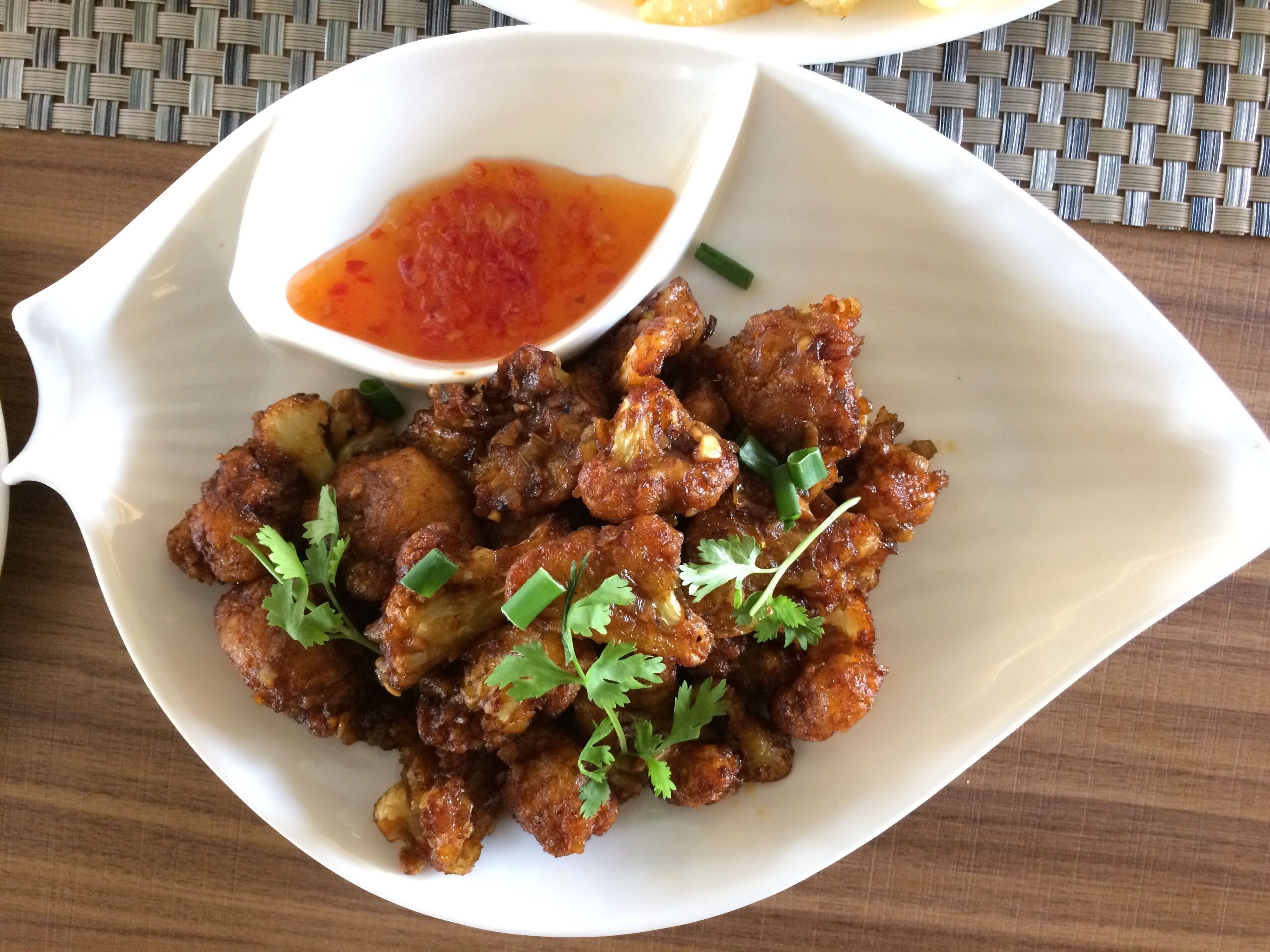 A white oval plate with fried chicken pieces garnished with green onions and cilantro, accompanied by a small bowl of red sauce.