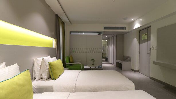 A hotel room with two beds, a green chair, a small table with a plant, and a bathroom visible through an open door.