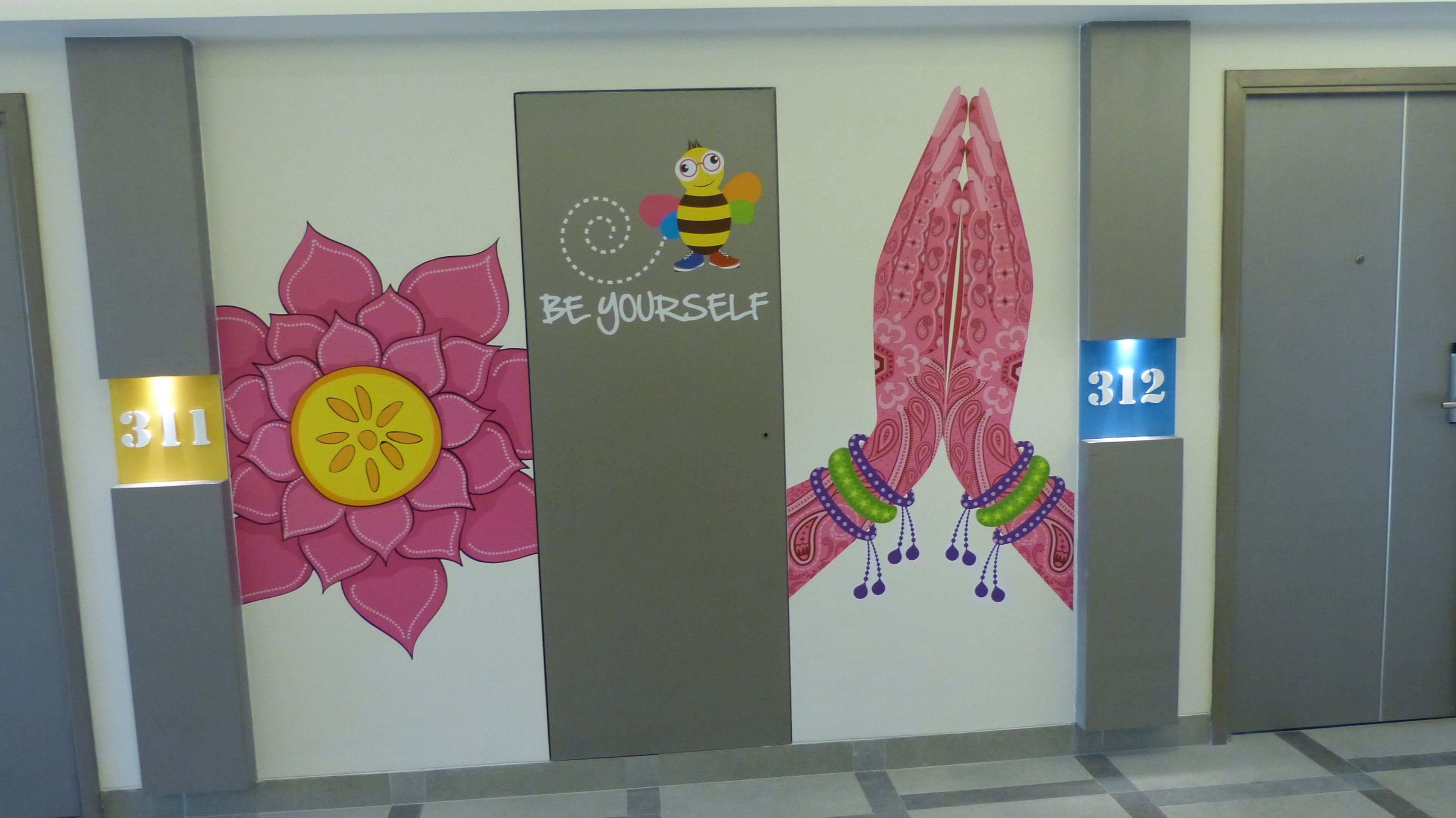 A hallway with a wall decorated with a large flower, a bee illustration, and a motivational phrase. There are two doors, one marked with the number 311 and the other with 312.