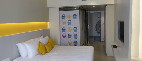 A modern hotel room with a large bed, white bedding, and yellow pillows. There is a wall-mounted TV and a door with colorful vehicle decals.