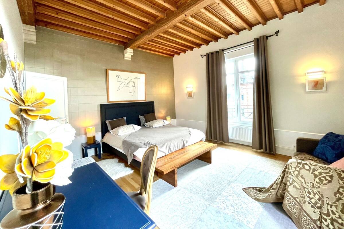A bedroom with a wooden ceiling, a bed with pillows, a bench, a desk with a vase of flowers, and a window with curtains.