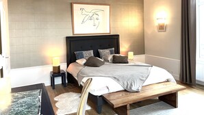 Ein Schlafzimmer mit einem großen Bett, einem Holzkopfstück, Nachttischlampen, einem gerahmten Kunstwerk und einem Fenster mit Vorhängen.