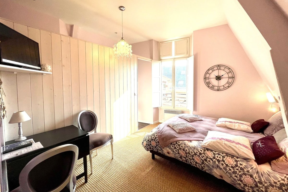 A bedroom with a bed, a desk, a chair, a chandelier, a clock, and a window.