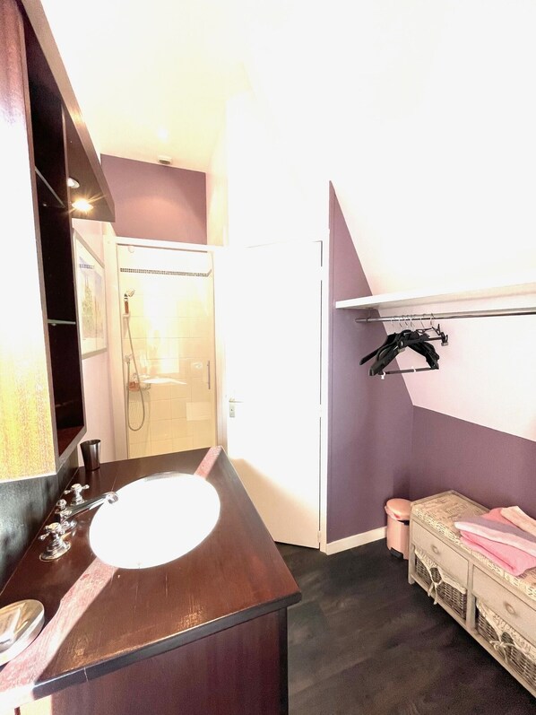 A bathroom with a wooden vanity, a white sink, a shower, and a small bedside table with a pink towel. - Maunoury Citybreak (Chartres)
