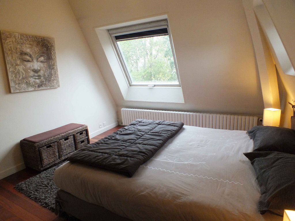 A bedroom with a large bed, a window, a radiator, a bedside table, and a wall-mounted artwork.