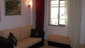 Une pièce avec un canapé beige, un petit pouf, un tableau au mur, une fenêtre avec des rideaux et une lampe.