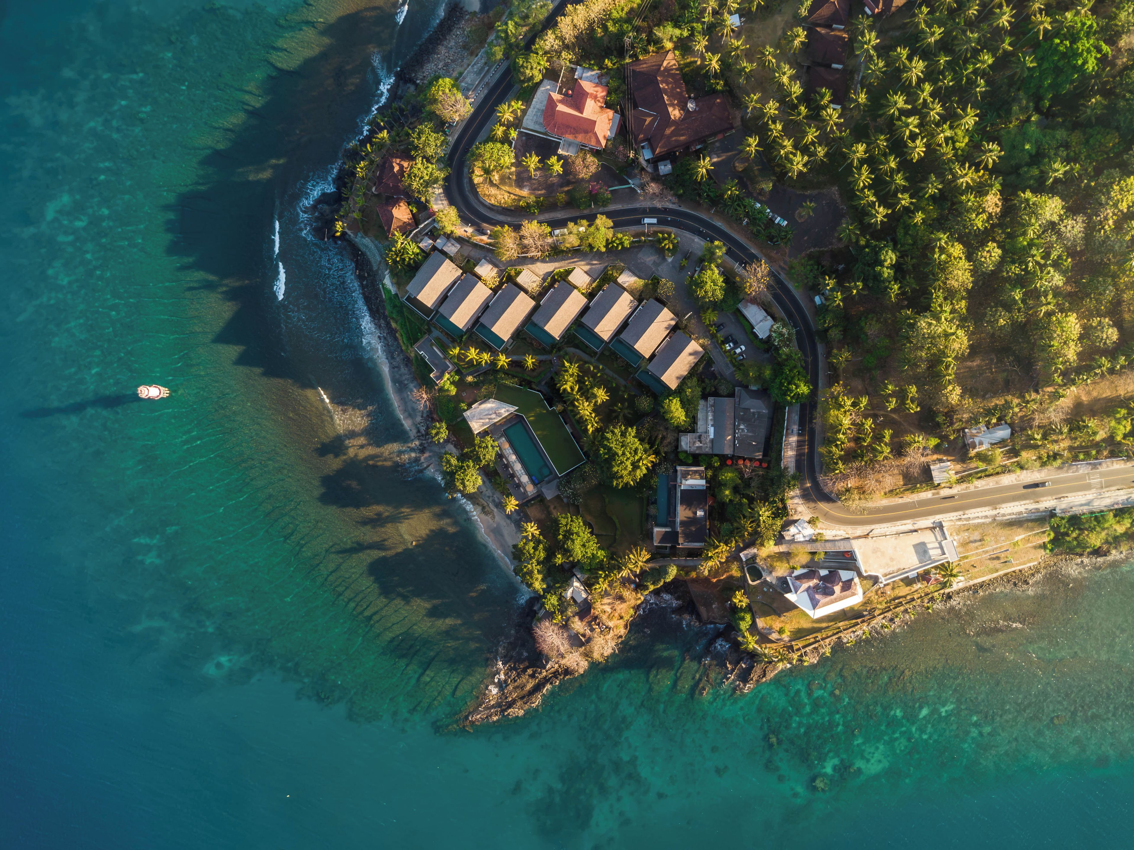 Vista aérea de uma propriedade costeira com uma estrada sinuosa, águas azuis cristalinas e uma pequena área de praia.