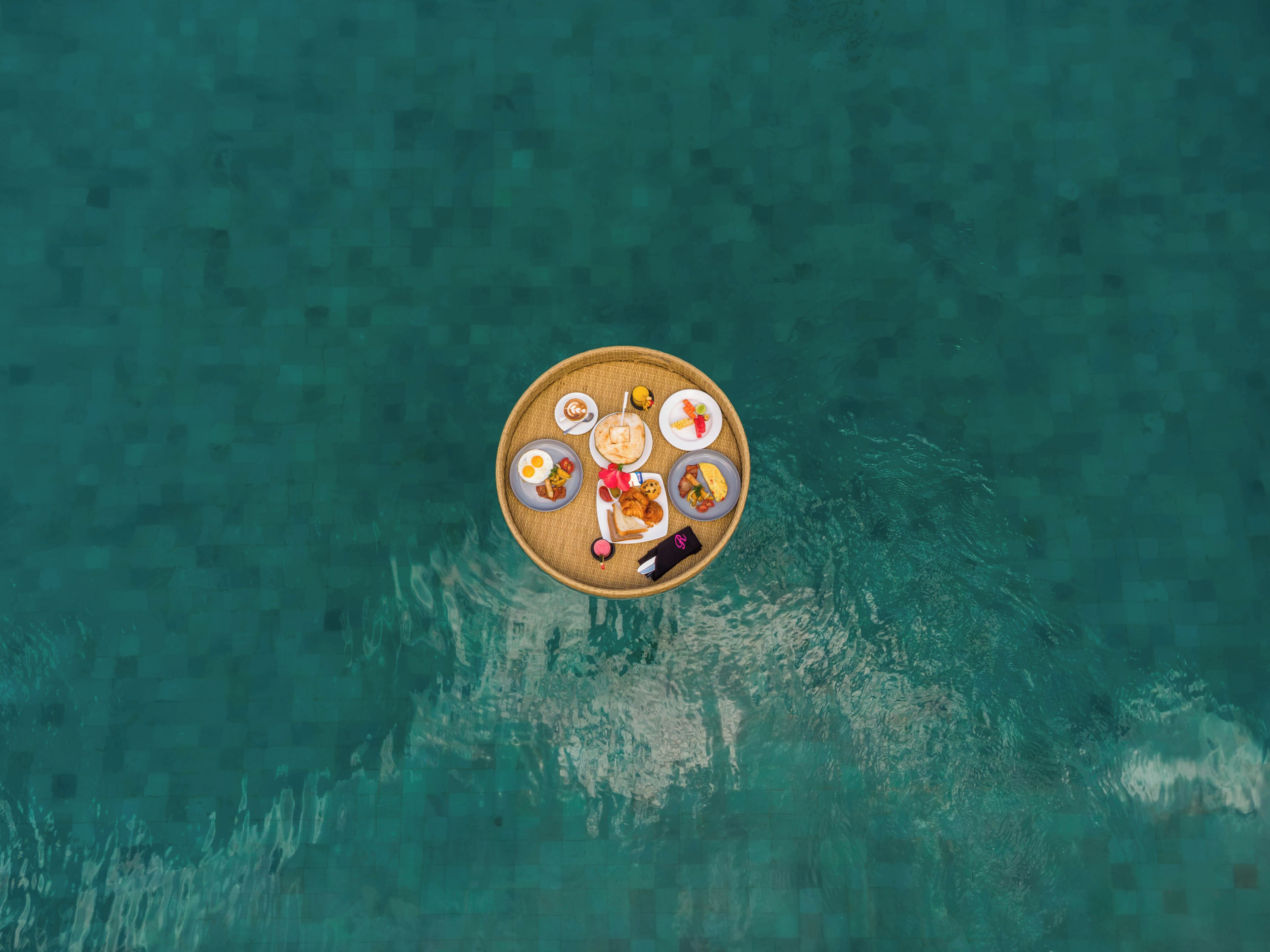 Vista aerea di una zona pranzo con un tavolo rotondo apparecchiato con diversi piatti, circondata da una piscina.
