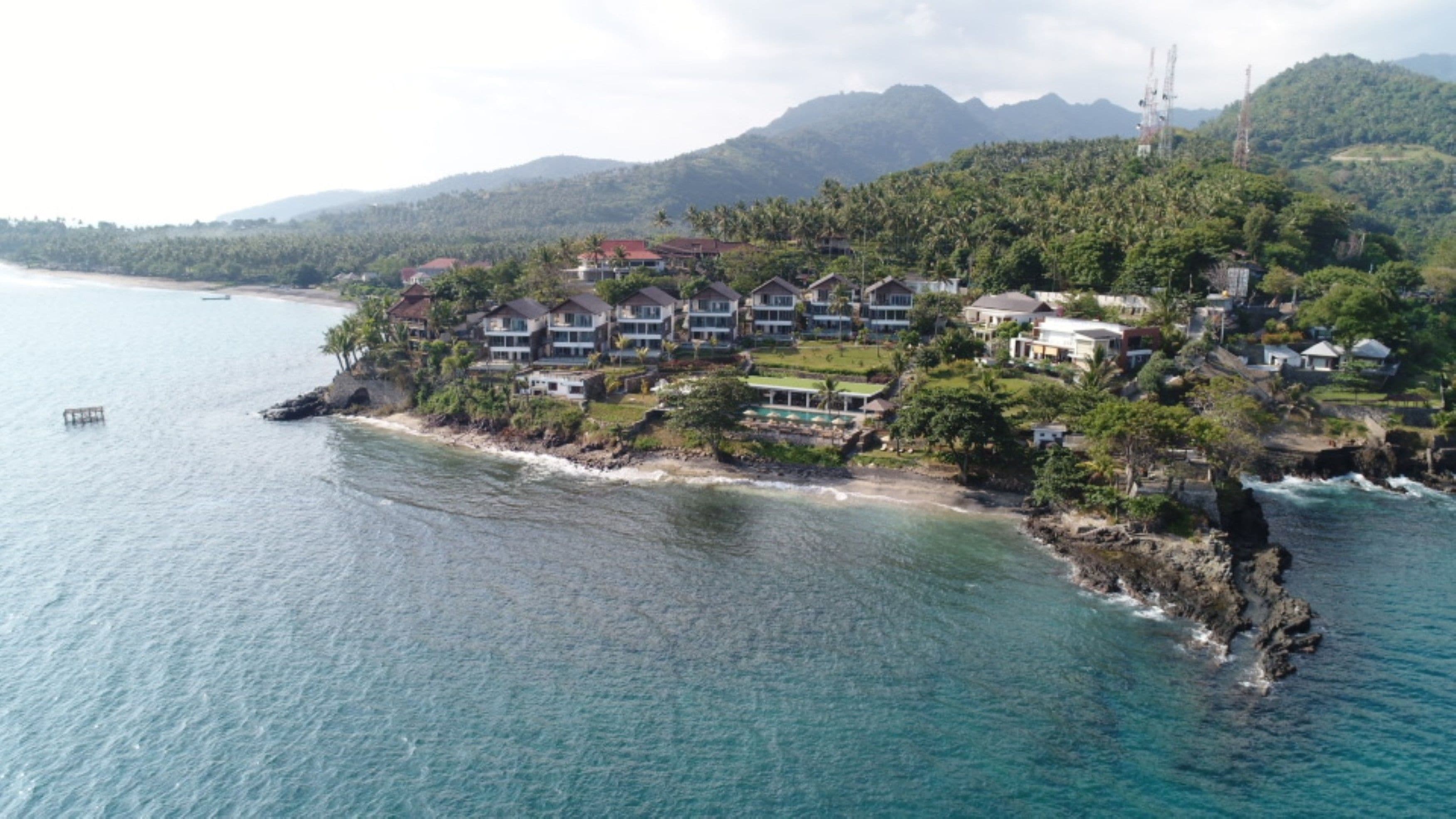 Ein Küstenresort mit mehreren Gebäuden, einem Strand und einer gebirgigen Kulisse.