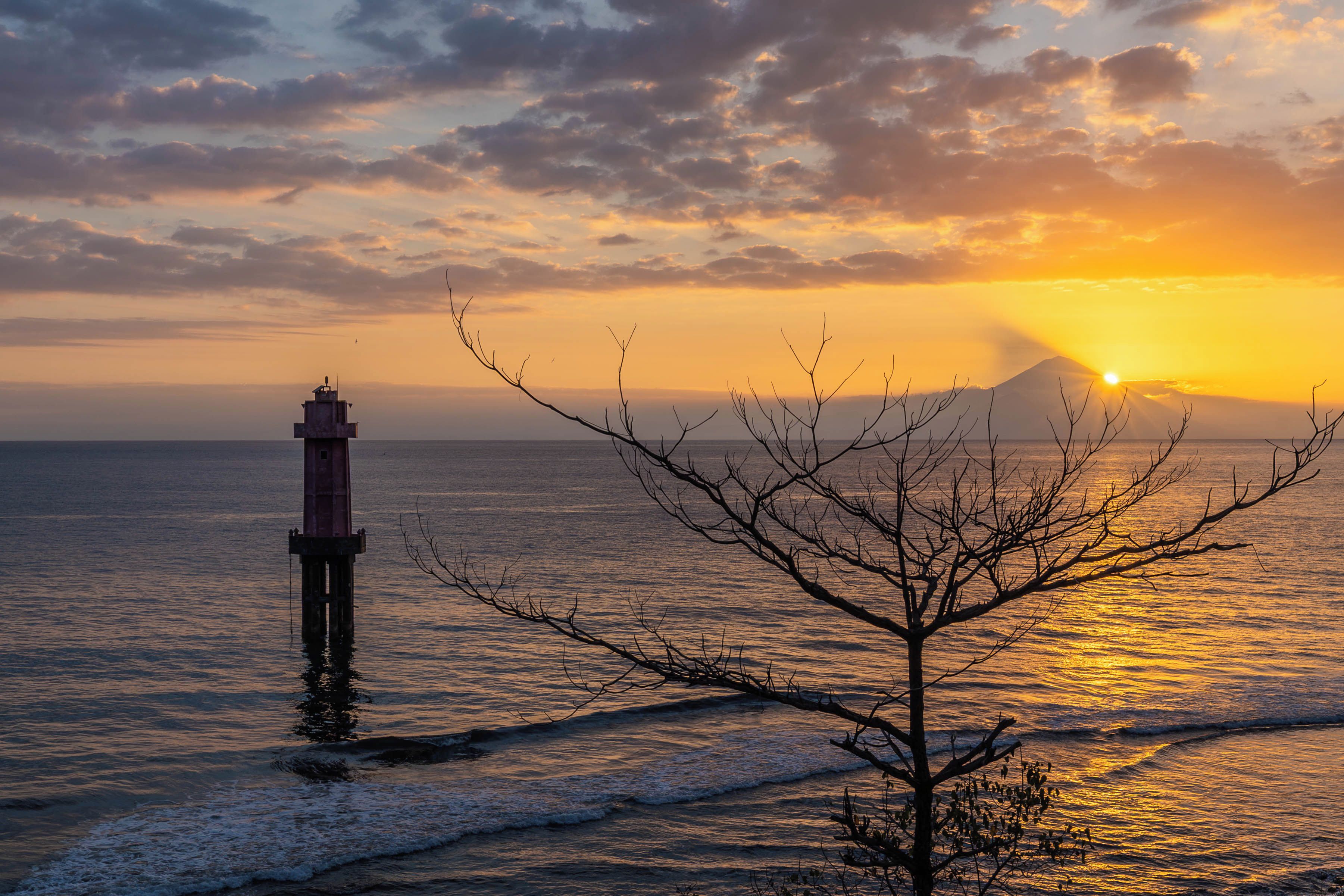 A lighthouse by the sea during sunset with waves approaching the shore.
