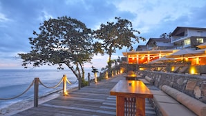 A wooden deck with a bench and cushioned seating, overlooking a beach and a building with outdoor lighting. - Rajavilla Lombok Resort (Senggigi)