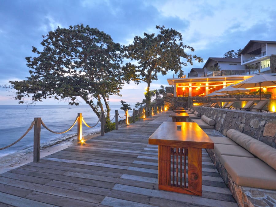 A wooden deck with a bench and cushioned seating, overlooking a beach and a building with outdoor lighting.