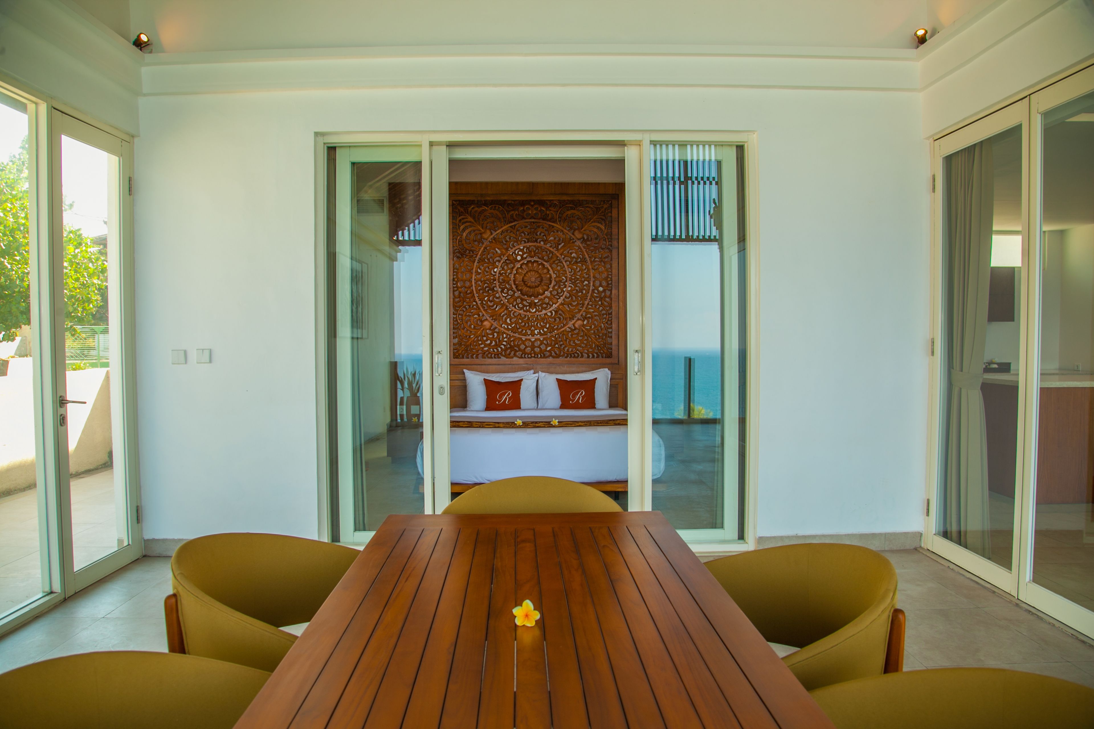 A room with a wooden table and yellow chairs, a bed with a patterned headboard, and a sliding glass door leading to a balcony.