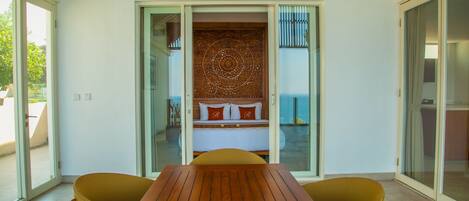 A room with a wooden table and yellow chairs, a bed with a patterned headboard, and a sliding glass door leading to a balcony.