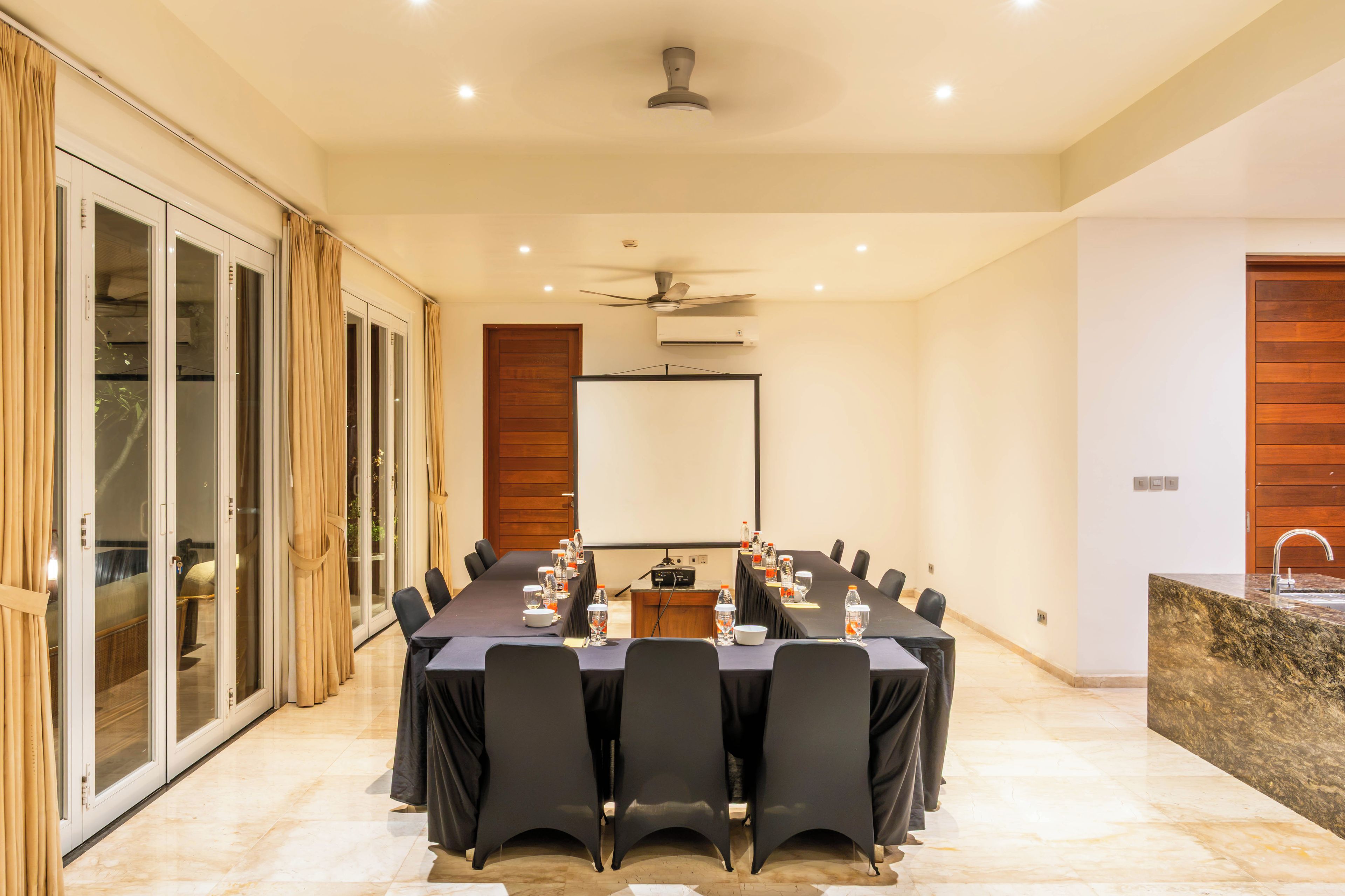 A conference room with a long table, chairs, a projector screen, and a ceiling fan.