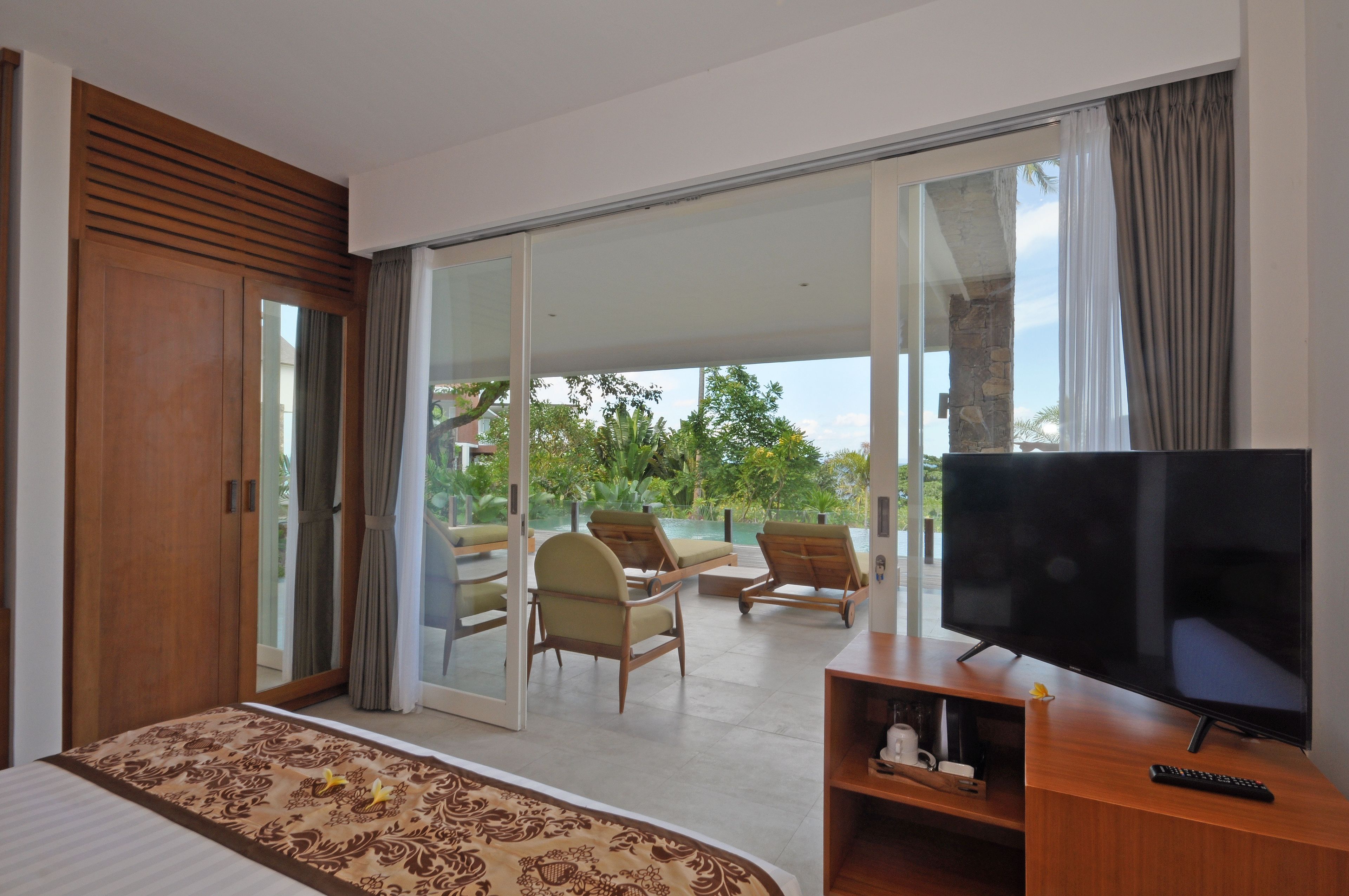 A bedroom with a bed, a TV, and a balcony with chairs and a view of greenery.