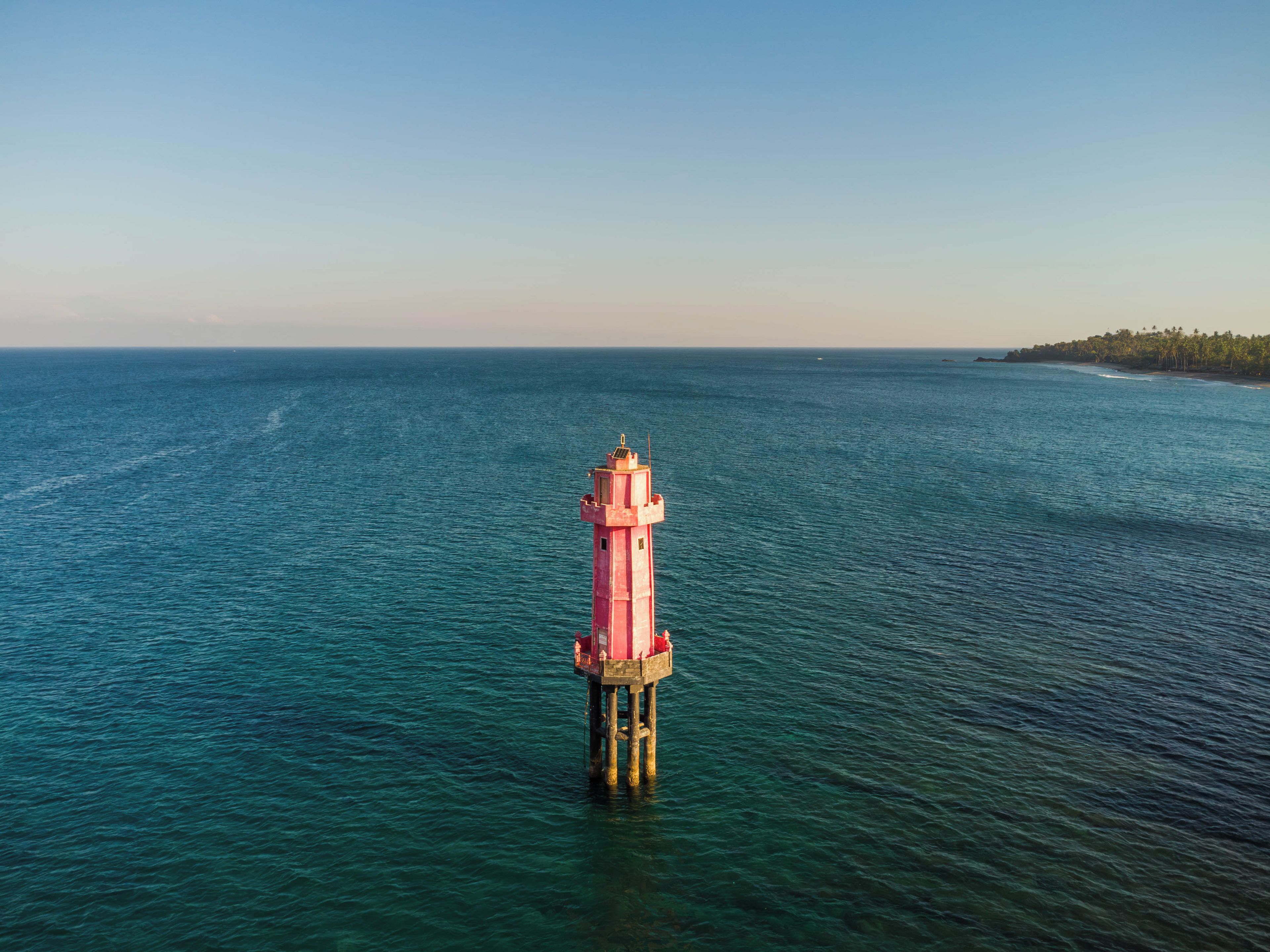 Un faro en un muelle que se adentra en un mar tranquilo, con un cielo despejado y una costa lejana con vegetación.