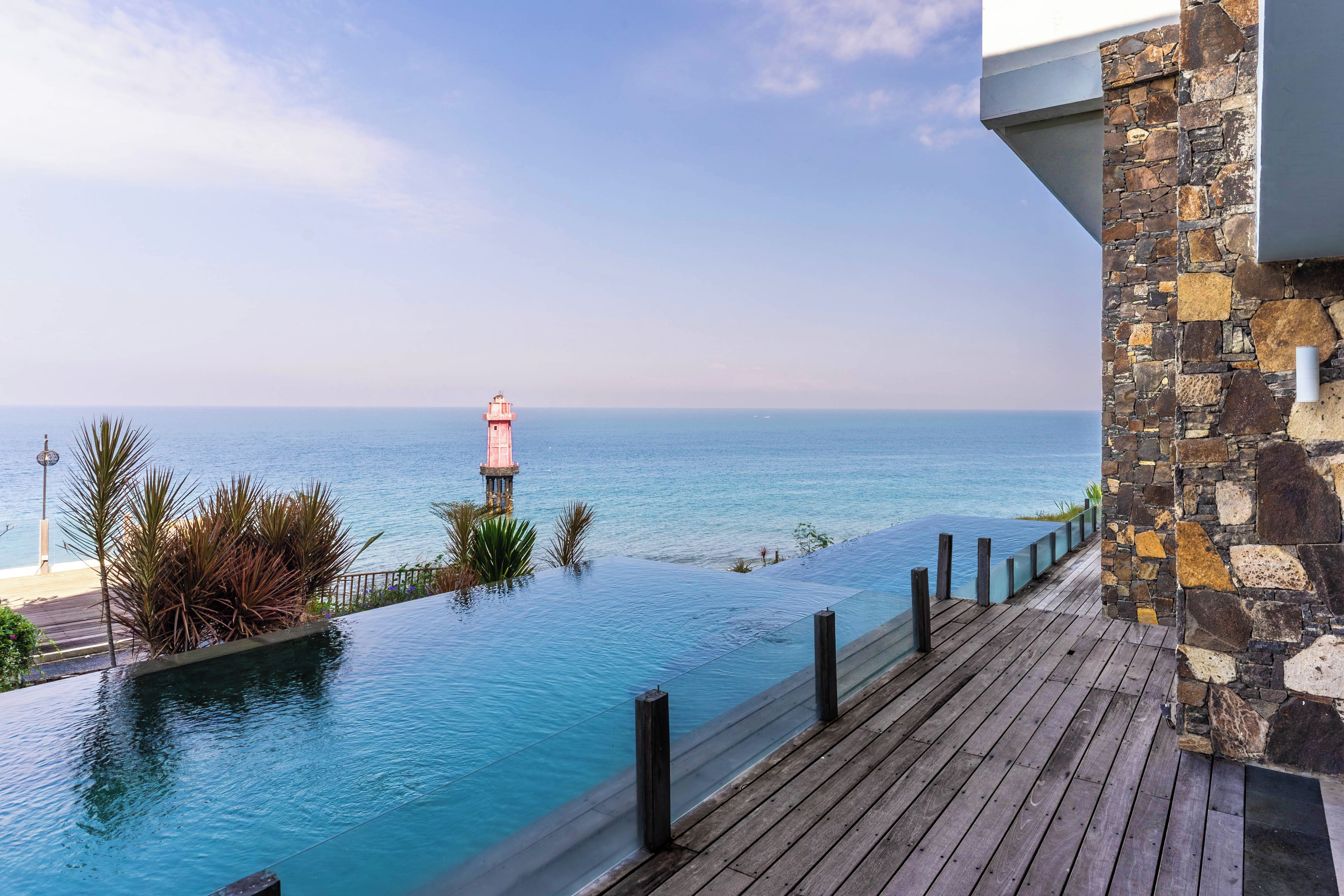 Una terraza de madera con vistas a un mar azul y despejado, con un faro a lo lejos.