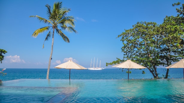 An infinity pool with clear blue water, palm trees, and umbrellas. - Rajavilla Lombok Resort (Senggigi)