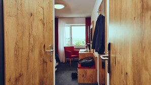 A hotel room with a wooden door, a red chair, a small table, a suitcase, and a view of the outdoors. - Die Burg (Donaueschingen)