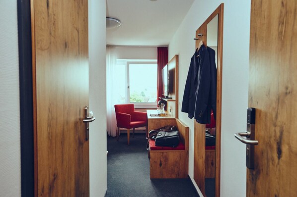 Habitación de hotel con puerta de madera, una silla roja, un escritorio con computadora, perchero y ventana con vista.