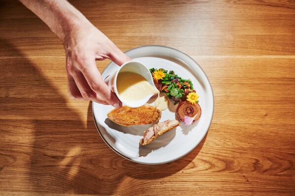 Un plato con un trozo de comida frita, una porción de carne y una pequeña taza de salsa sobre una mesa de madera.