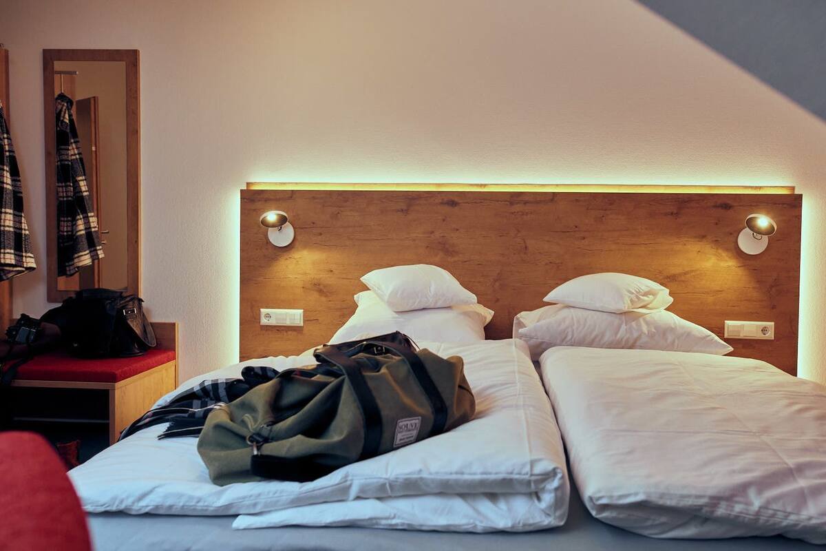 A hotel room with a wooden headboard, two beds, a red bench, and a coat rack with a plaid jacket.