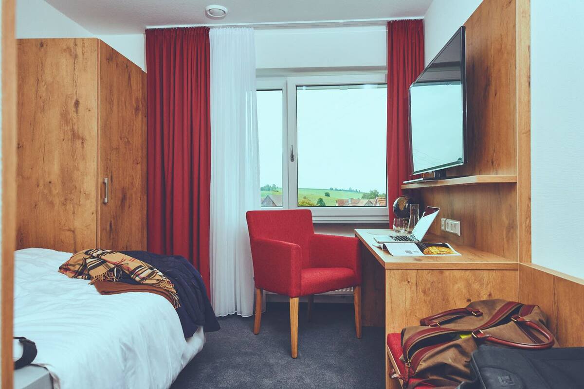 A hotel room with a bed, a desk with a laptop, a chair, and a TV on the wall.