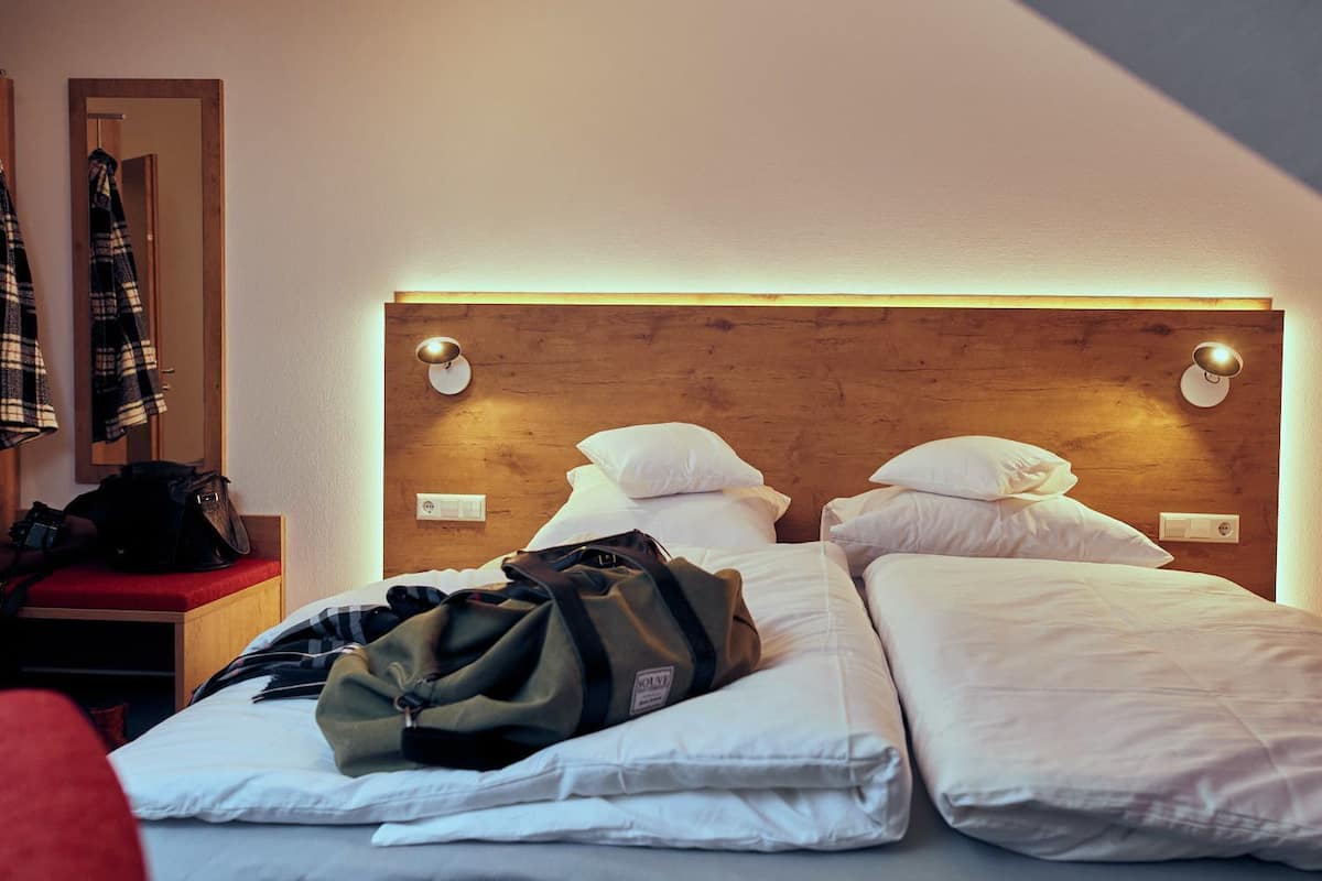 A hotel room with a wooden headboard, two beds, a red bench, and a coat rack with a plaid jacket.