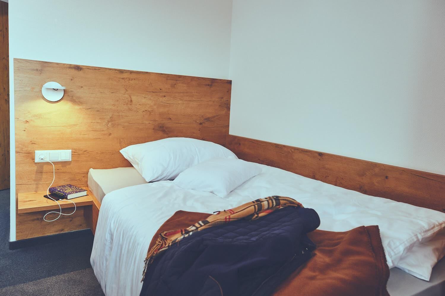 A single bed with white bedding, a wooden headboard, a bedside table with a book and a power outlet, and a wall-mounted light.