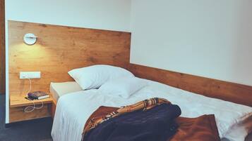 A single bed with white bedding, a wooden headboard, a bedside table with a book and a power outlet, and a wall-mounted light.