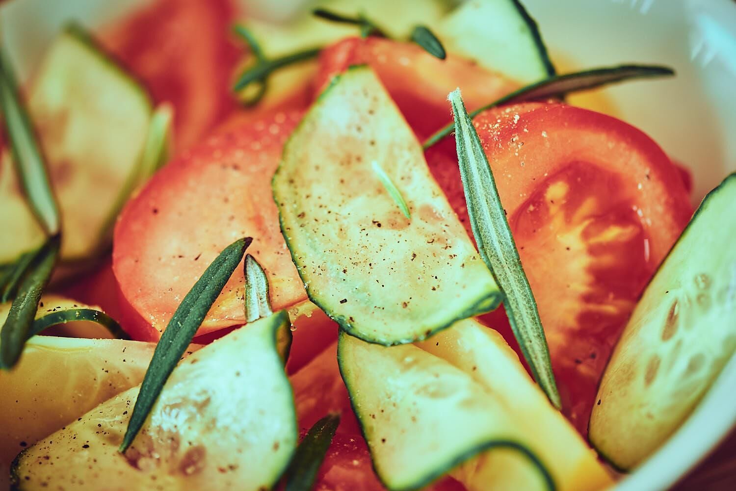 A bowl of sliced tomatoes and cucumbers with rosemary sprigs.
