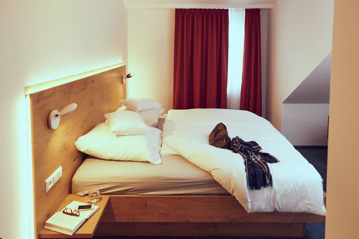 A neatly made bed with pillows and a blanket, a bedside table with a book and glasses, and a red curtain in the background.