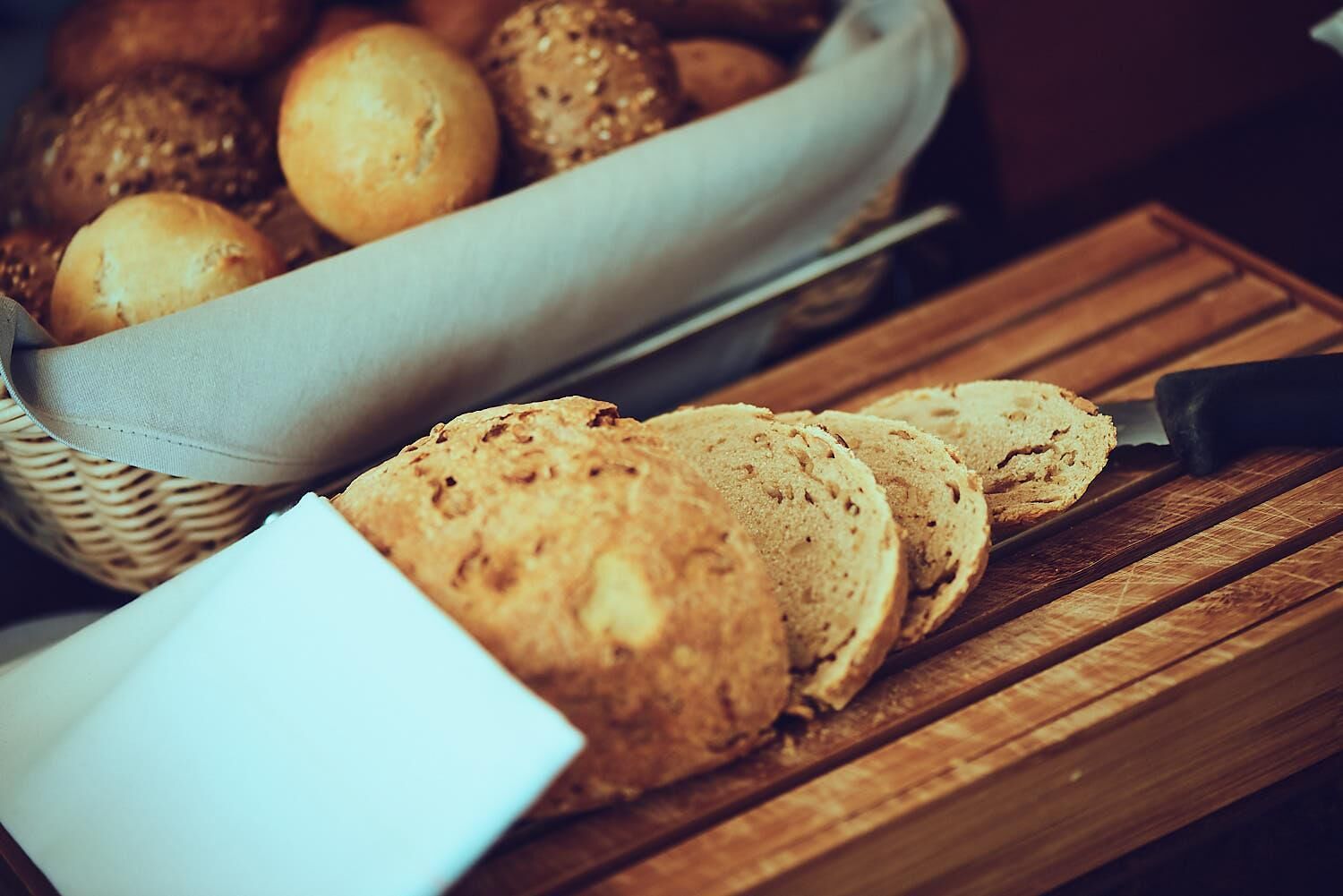 Brotscheiben auf einem Holzschnittbrett, ein Korb mit runden Brötchen und eine weiße Serviette.